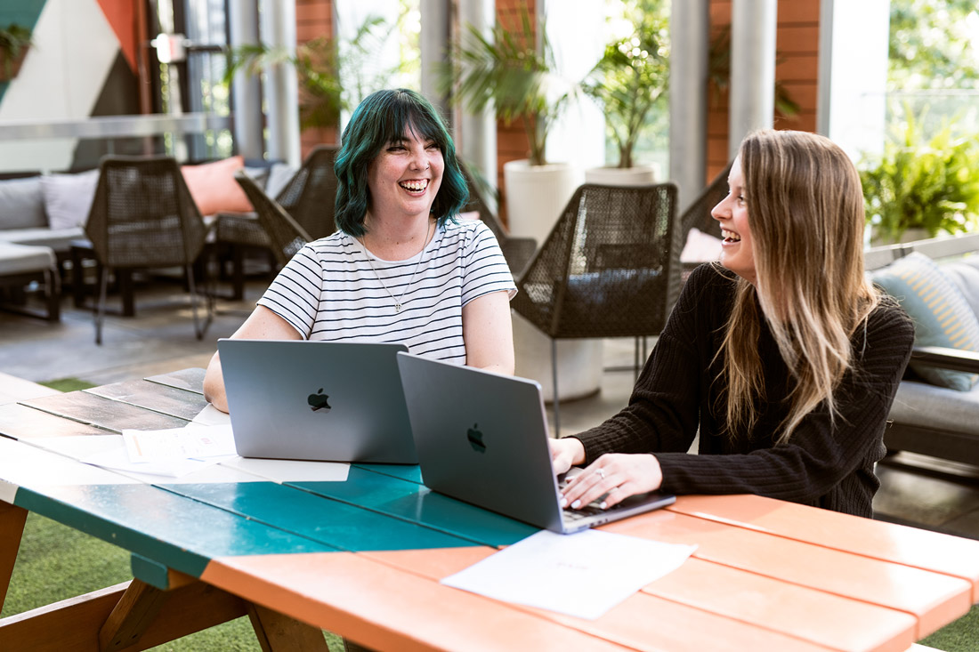 Conheça os Especialistas por Trás de Nossa Plataforma de Faturamento - Equipe Invoice Home Dois membros da equipe da Invoice Home discutindo o trabalho em seus laptops em um escritório casual ao ar livre.